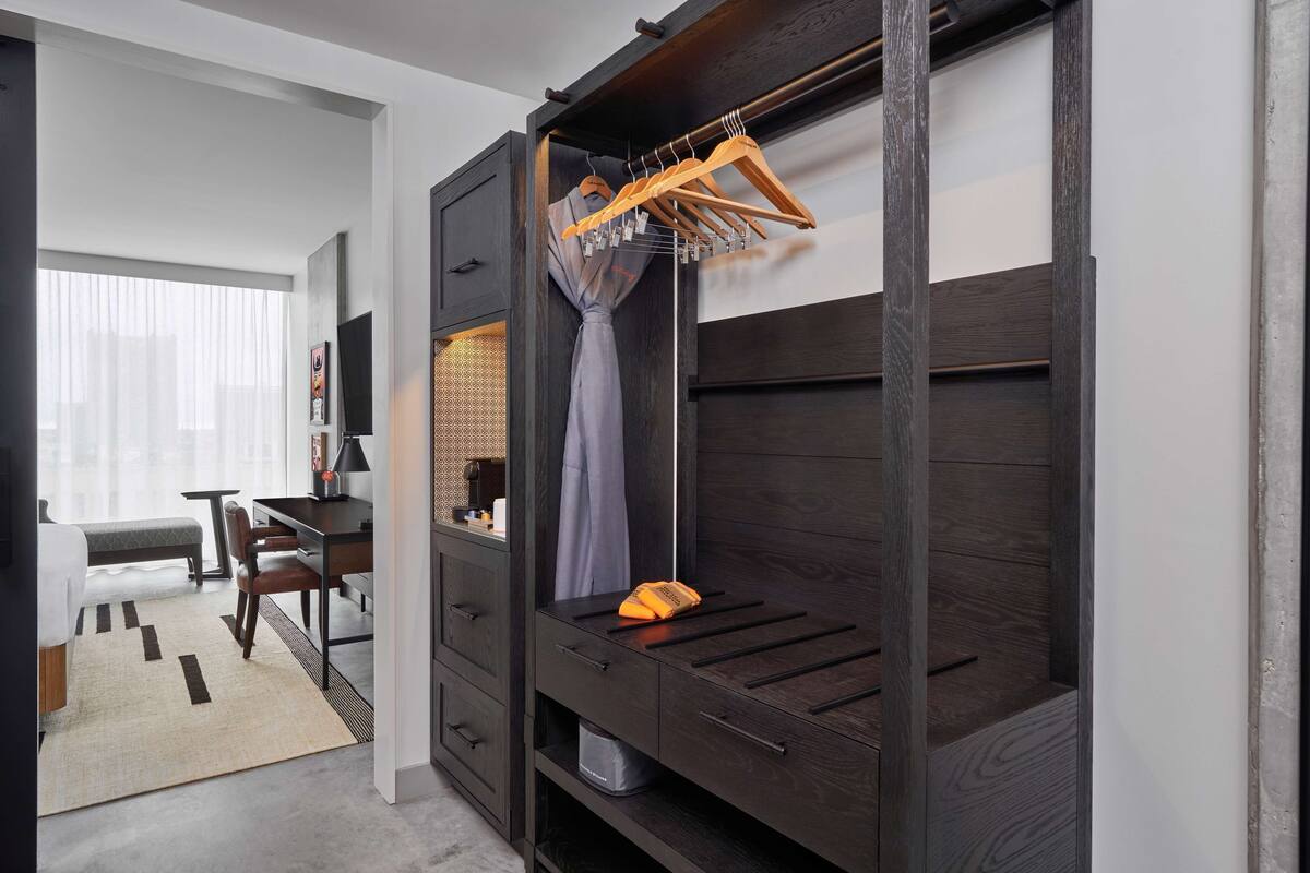 A modern hotel room with a dark wood wardrobe, a desk with a chair, a bed, and a large window with sheer curtains.