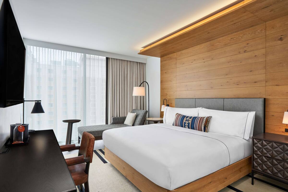 A modern hotel room with a large bed, a desk, a chair, a small table, and a lamp.