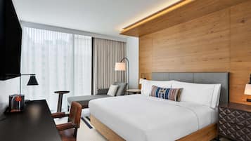 A modern hotel room with a large bed, a desk, a chair, a small table, and a lamp.