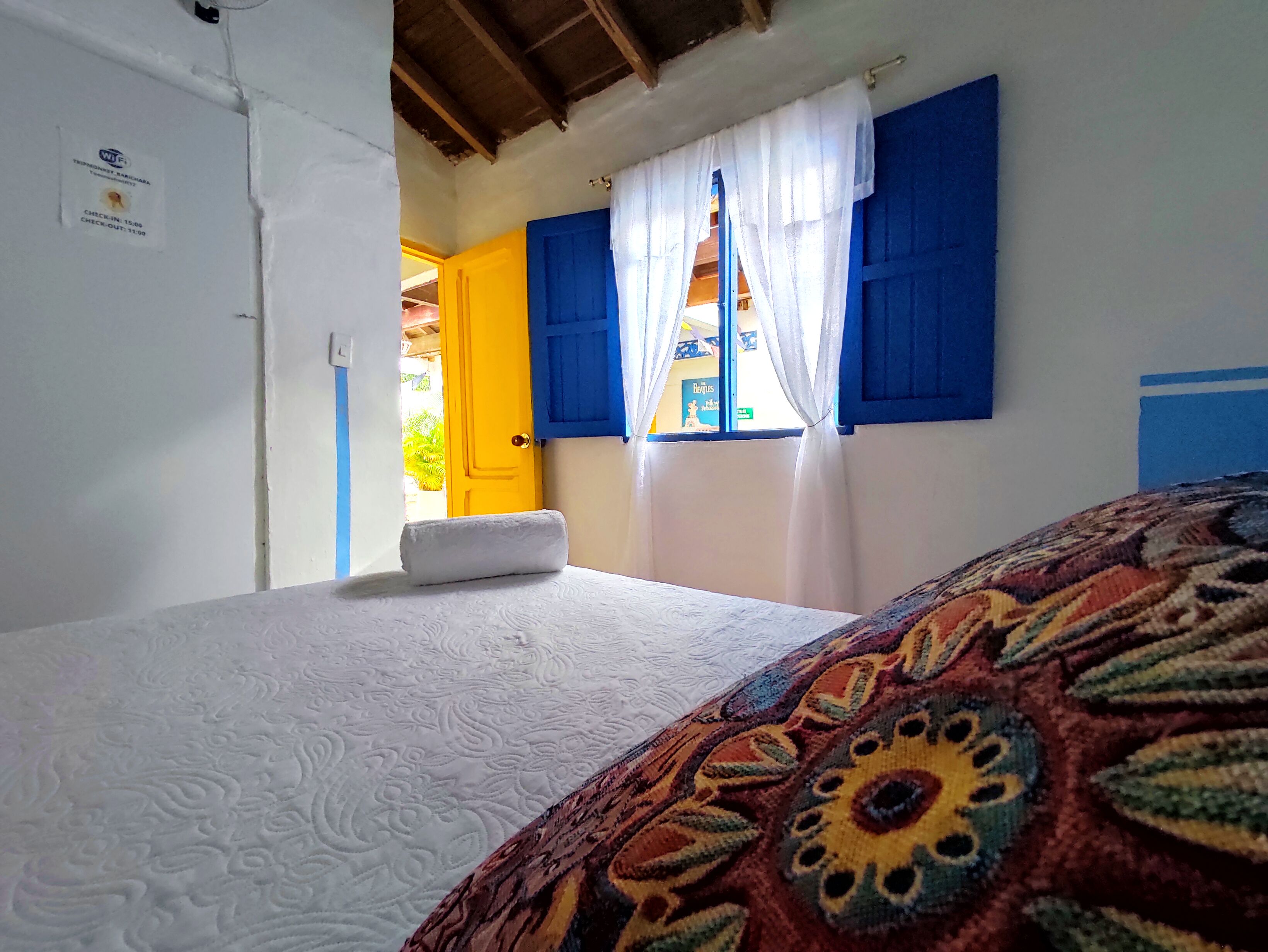 Una habitación con cama, ventana con contraventanas azules, una puerta y un cartel de recepción.
