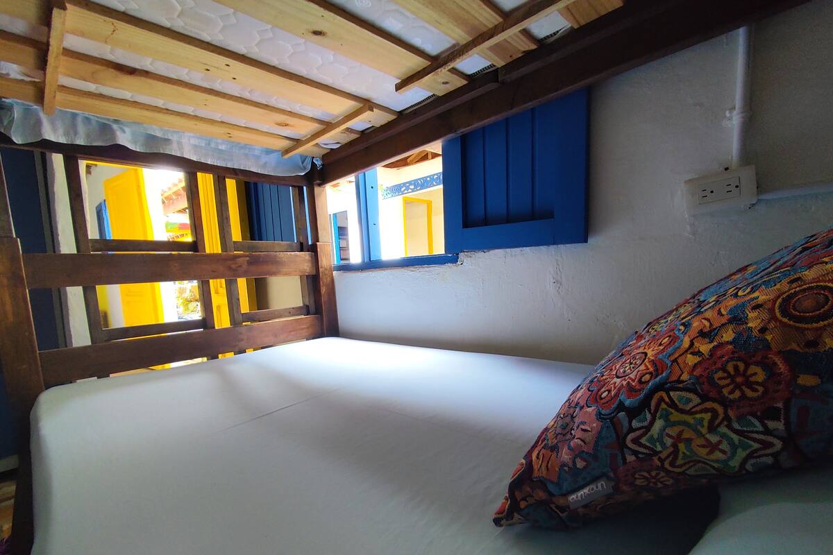 Une chambre avec un lit, des poutres en bois, une fenêtre avec des volets bleus et un coussin à motifs.