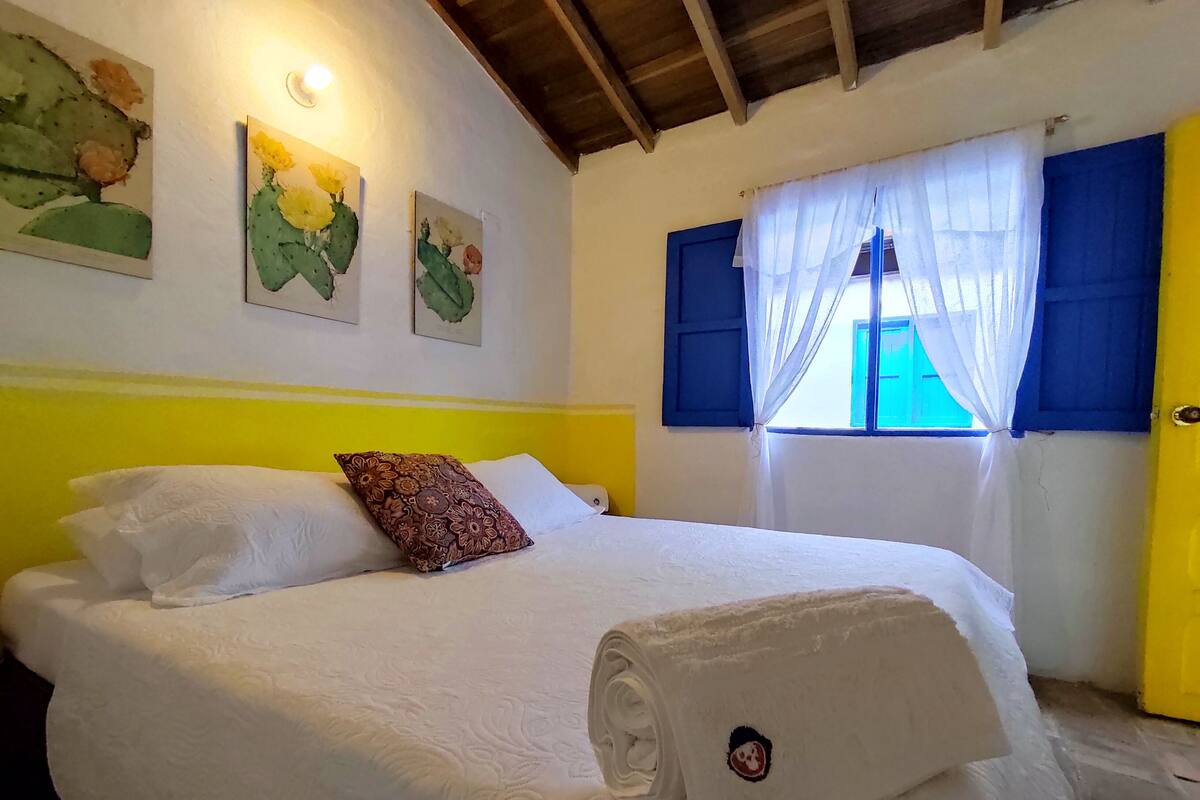Un lit bien fait, avec du linge de lit blanc et une serviette enroulée, un mur jaune et trois tableaux représentant des cactus.