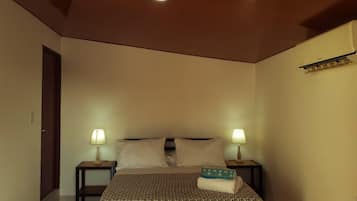 A bedroom with a bed, two bedside tables with lamps, a wooden ceiling, and an air conditioning unit on the wall.