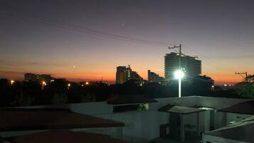 A rooftop with a view of a city skyline at dusk, featuring a prominent high-rise building and several smaller structures.