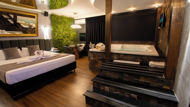 A modern hotel room with a bed, a seating area, a green wall, a fireplace, and a TV.