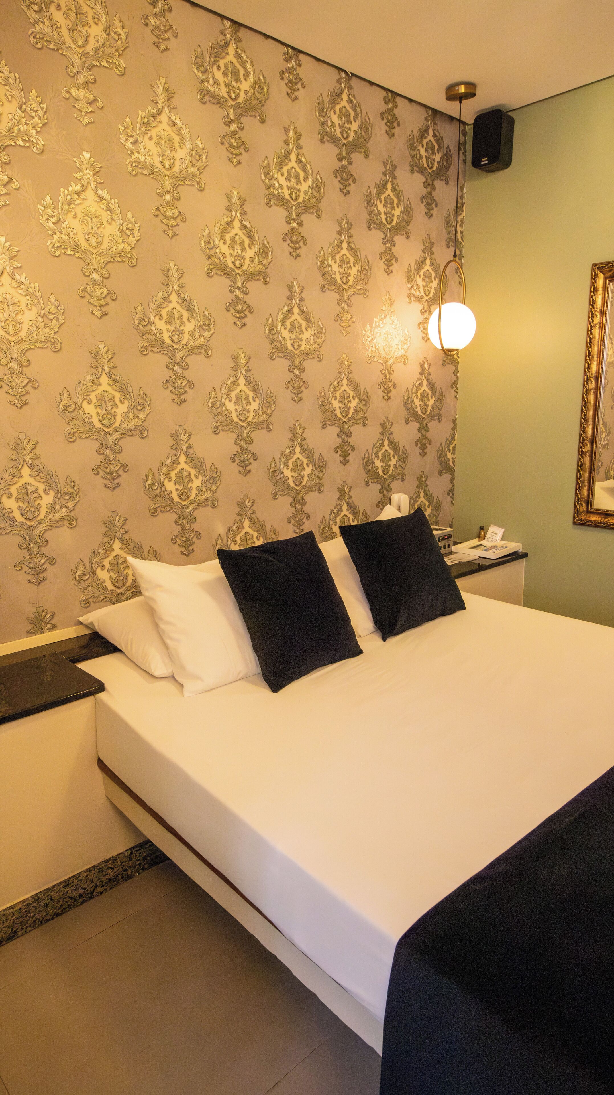 Une chambre d’hôtel avec un grand lit, deux oreillers noirs, une table de chevet avec un téléphone, une lampe fixée au mur et un tableau encadré.