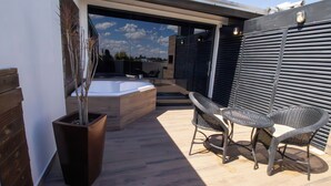 A modern outdoor patio with a hot tub, two wicker chairs, and a wooden planter with a small tree.
