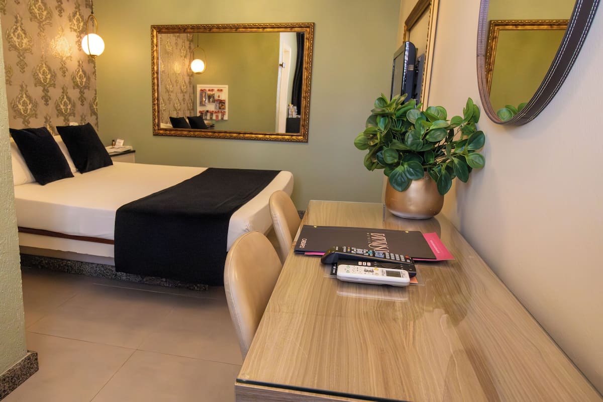 Ein Hotelzimmer mit einem Bett, einem Nachttisch, zwei Stühlen, einem Schreibtisch, einem Spiegel, einer Topfpflanze und einer Wand mit strukturierter Tapete.