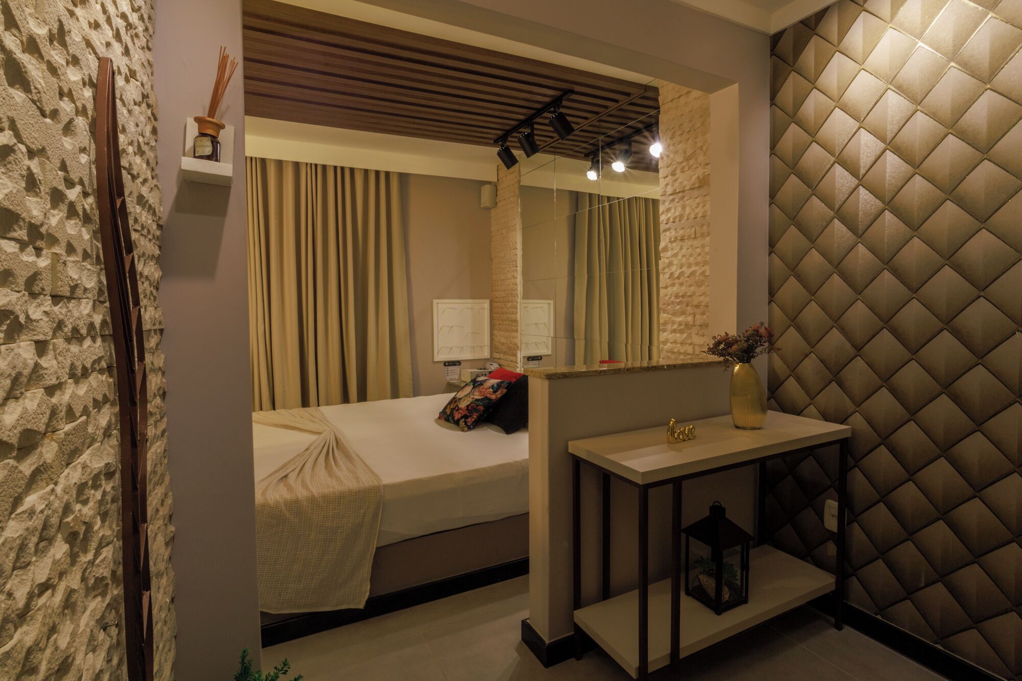 A hotel room with a bed, a bedside table with a lamp, a mirror, and a textured wall.