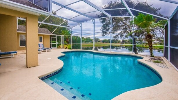 A swimming pool with a curved edge, surrounded by a covered area with a glass roof, and a view of a lake and trees in the background.