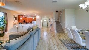 A modern living room with a sofa, dining table, and chairs. There is a kitchen in the background with cabinets and appliances. A staircase is visible to the right. - Ultimate Champions Gate Resort 8 Bedroom 5 Bath (Davenport)