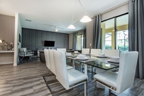 A modern dining area with a glass table and white chairs, a flat-screen TV, and a view through large windows. - 6 Bedroom 5 Bathroom in Solterra Resort 5425sc (Davenport)