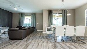 A modern living room with a sofa, dining table, and chairs, a ceiling fan, and large windows with curtains. - 6 Bedroom 5 Bathroom in Solterra Resort 5425sc (Davenport)