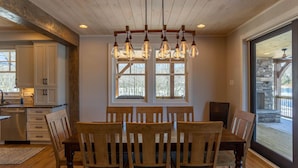 A dining area with a wooden table and chairs, a chandelier, and large windows with a view of the outdoors. - Lakeshore Retreat- new construction in Sweetgrass, right on the lake (Blowing Rock)