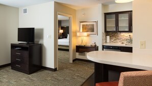 Ein Hotelzimmer mit Flachbildfernseher, Schreibtisch mit Lampe, einer kleinen Küchenzeile und einem Bett im Hintergrund.