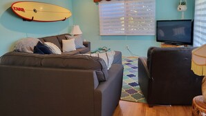 A living room with a sofa, a television, a ceiling fan, a surfboard, and a window with blinds. - The Williams Cottage (Carlsbad)