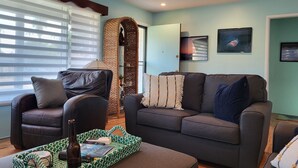 A living room with a sofa, armchair, and a coffee table with a bottle and books. - The Williams Cottage (Carlsbad)