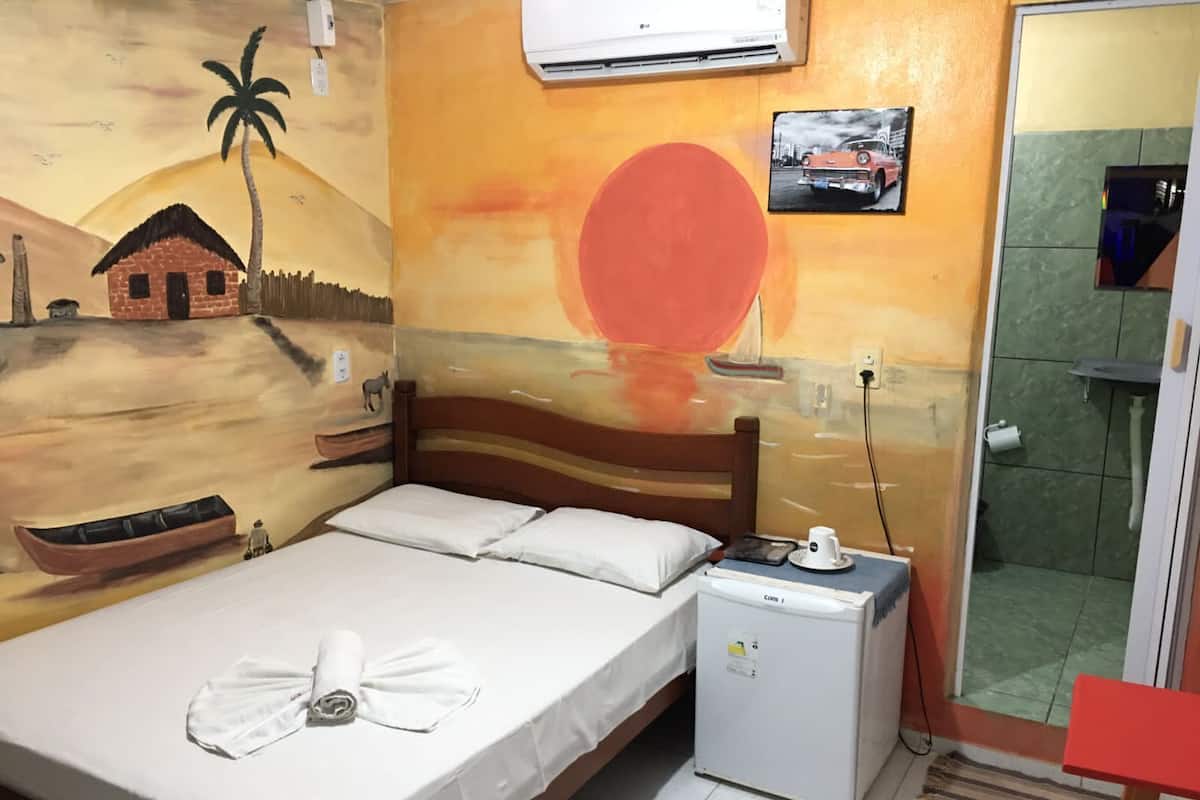 Quarto com cama, um pequeno frigorífico e um mural na parede retratando um cenário de pôr do sol.