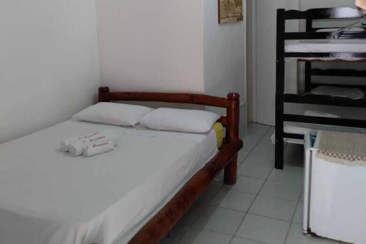 Uma cama de solteiro com estrutura de madeira e roupa de cama branca, uma pequena mesa com toalha xadrez e uma cama beliche com estrutura preta num quarto com paredes brancas e chão de azulejos.