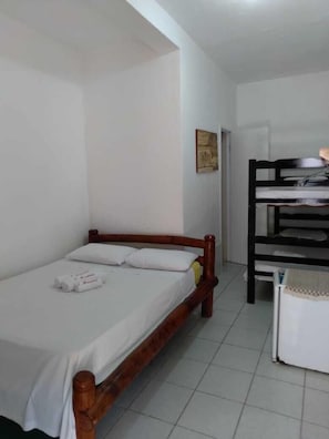A single bed with a wooden frame and white bedding, a small table with a checkered tablecloth, and a bunk bed with a black frame in a room with white walls and tiled flooring. - Pousada Casa Beneventi Jeri (Jijoca de Jericoacoara)