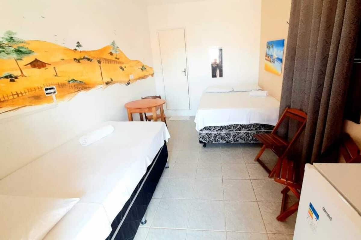 Quarto de hotel com duas camas, uma mesinha, uma cadeira, um refrigerador e um mural na parede.