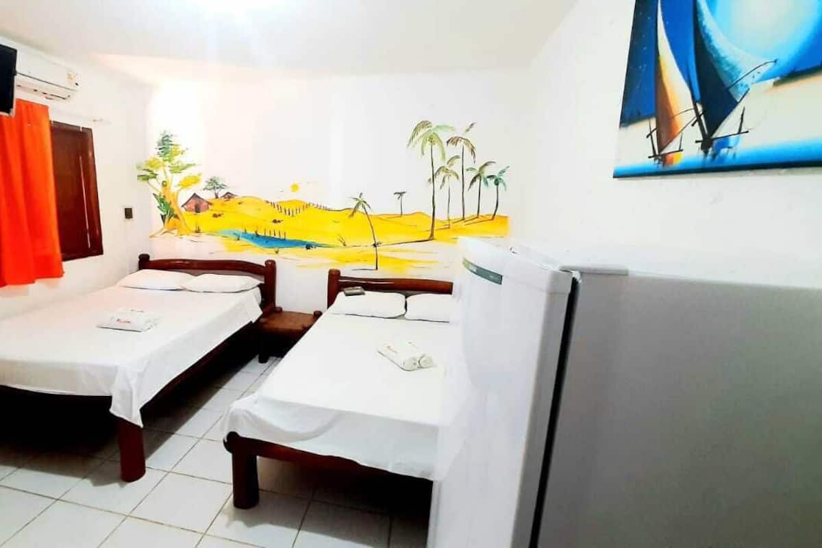 Quarto com duas camas de solteiro, uma TV fixada na parede, um frigorífico e um mural com uma paisagem de praia.