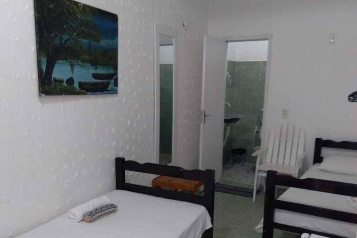 Quarto pequeno e simples com duas camas de solteiro, uma mesinha e um refrigerador.