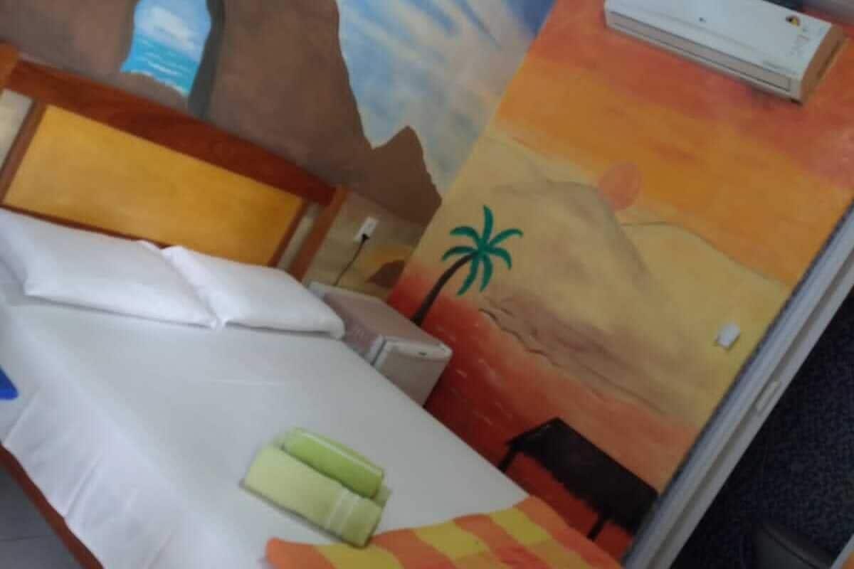 Cama com lençóis brancos, cabeceira de madeira e travesseiro colorido listrado. Há um ar condicionado fixado na parede e uma pintura de uma palmeira.