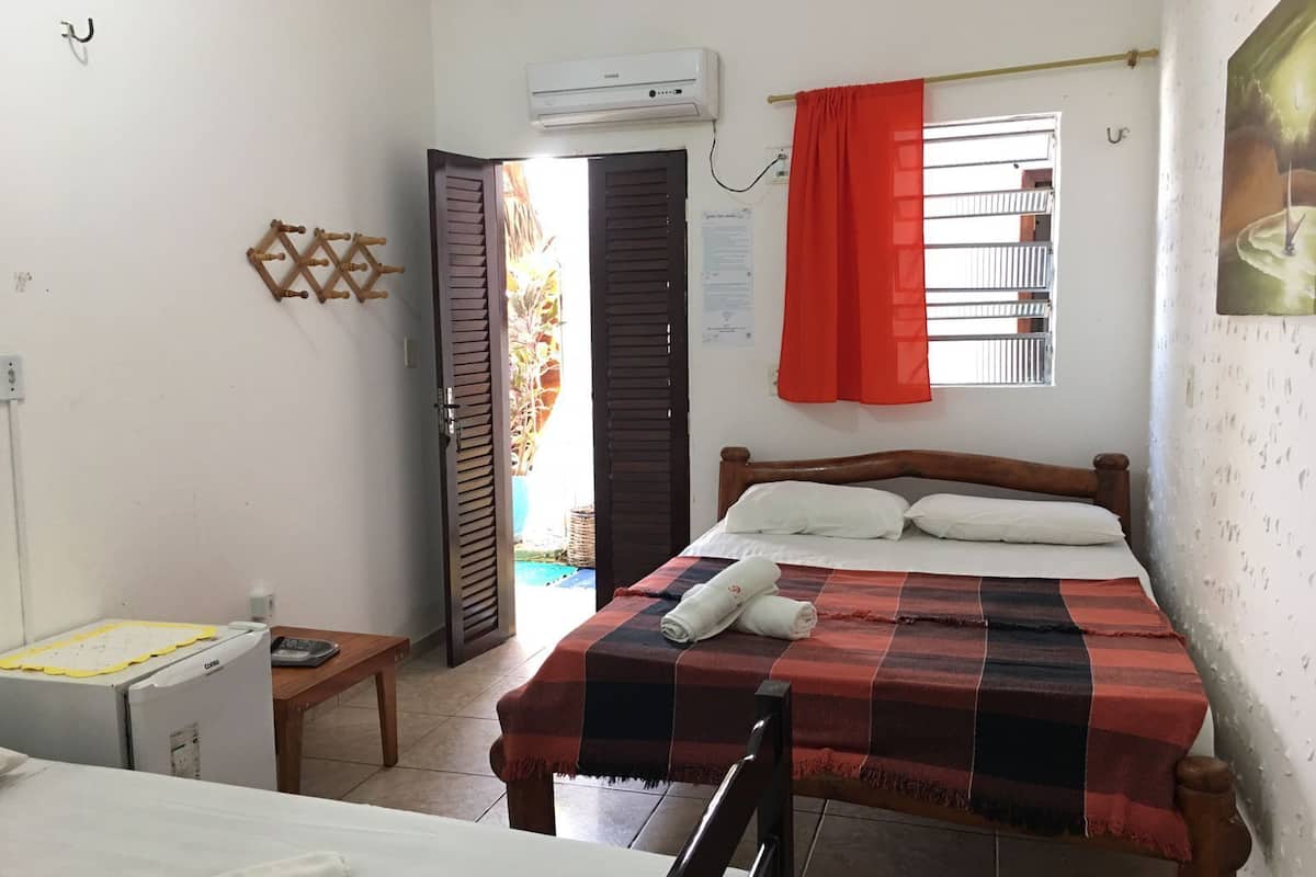 Quarto com cama, uma mesinha, uma cadeira, aparelho de ar condicionado, janela com cortina vermelha e porta que dá para o exterior.
