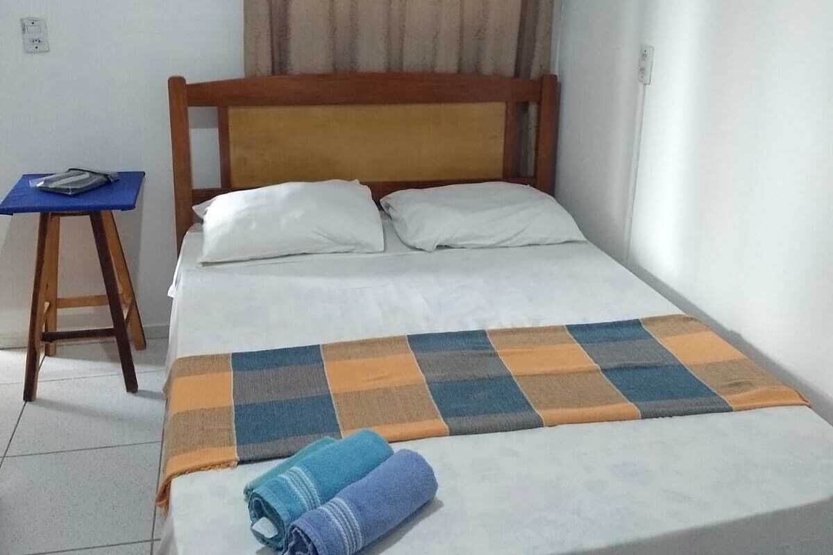 Uma cama de solteiro com cabeceira de madeira, manta xadrez e duas toalhas enroladas.