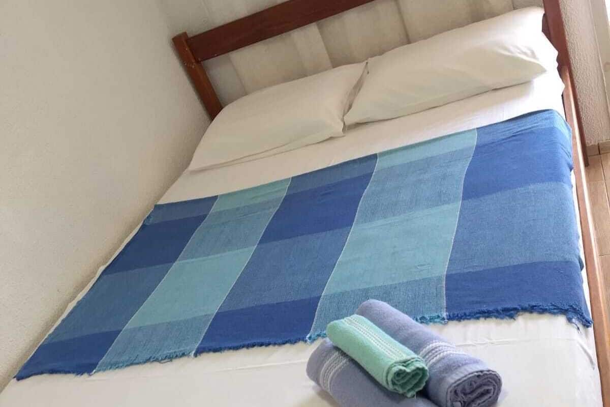 Uma cama com manta azul e branca xadrez, almofadas brancas e toalhas enroladas sobre a cama.