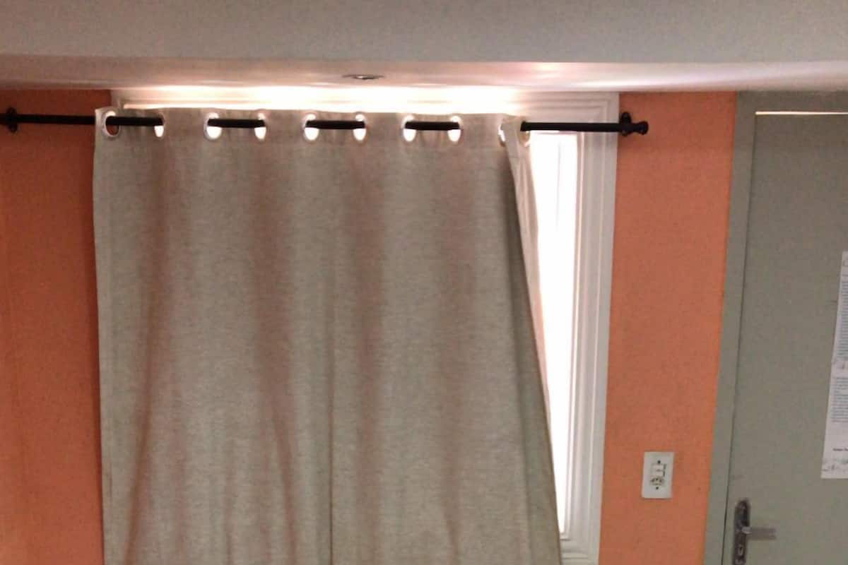 Uma cama de solteiro com colcha branca e duas almofadas, uma toalha verde e uma janela com cortinas.
