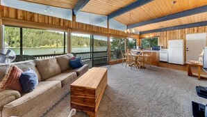 Un'ampia zona giorno con soffitto in legno, ampie finestre che si affacciano su una foresta, un divano, un tavolino da caffè in legno e un angolo cottura con frigorifero e piano cottura.