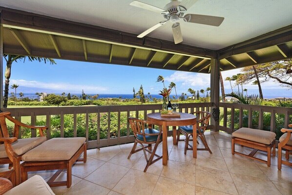Een overdekt terras met een tafel, stoelen, een plafondventilator en uitzicht op zee en groen.