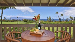 A round wooden table with a bottle of wine, a glass, and a fruit platter on a balcony overlooking a golf course and ocean.