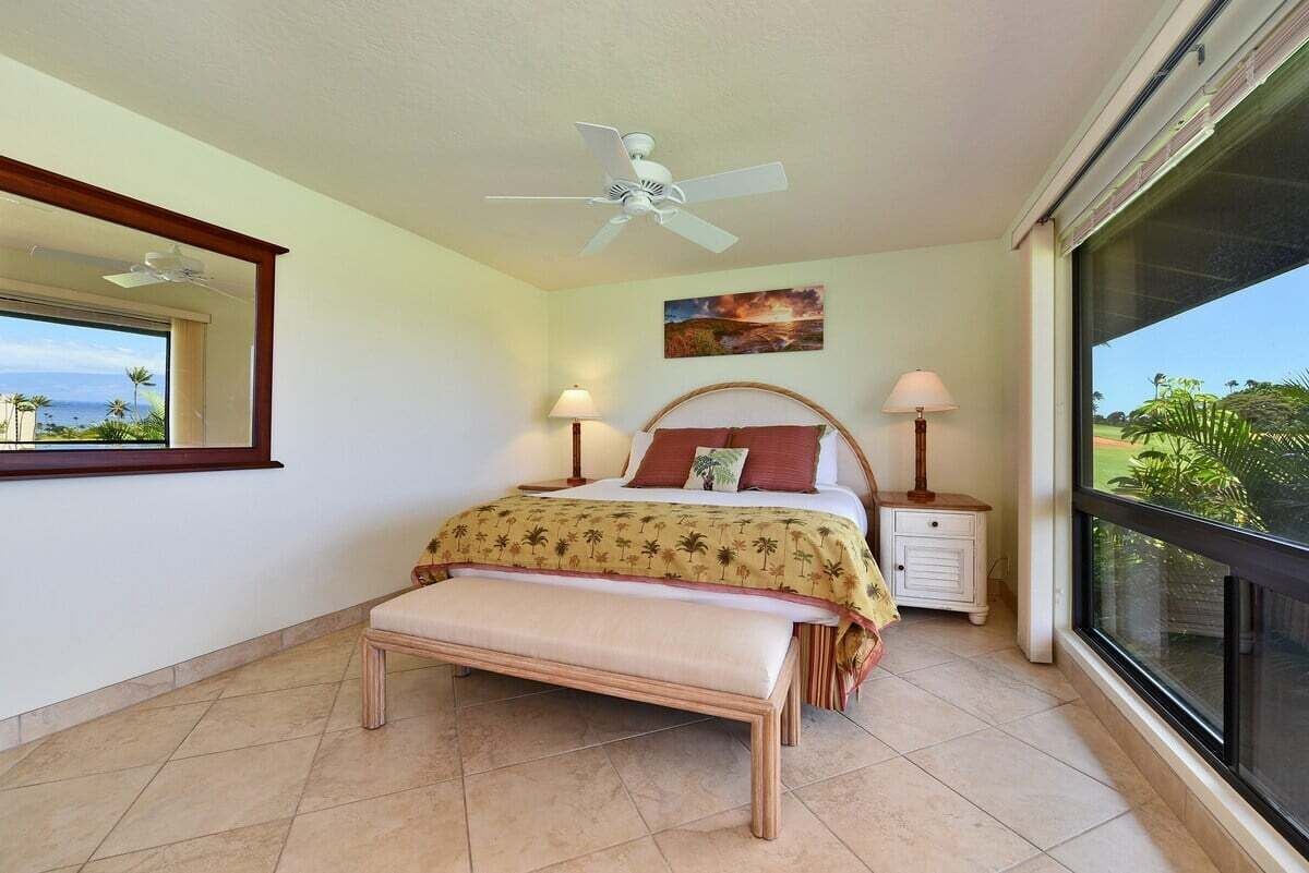 Un dormitorio con cama, banca, ventilador de techo, lámparas de noche y un ventanal con vista al océano.