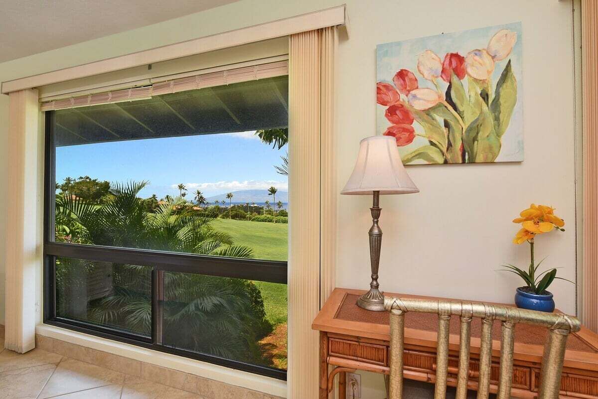 A room with a large window offering a view of a golf course and palm trees. There is a wooden desk with a lamp and a potted plant.
