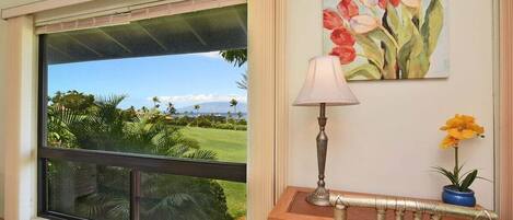 A room with a large window offering a view of a golf course and palm trees. There is a wooden desk with a lamp and a potted plant.