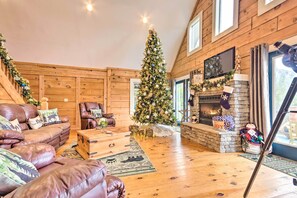 Interior acolhedor de uma cabana de madeira com árvore de Natal, sofás de couro, lareira de pedra e televisão na parede.