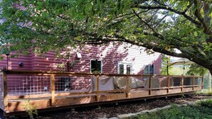 A wooden deck with a metal railing, a pink house in the background, and a tree providing shade. - Relax on the Ranch…..HOT TUB, SHEEP (Sultan)