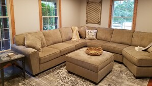 A L-shaped sectional sofa with a matching ottoman, a small table with a patterned table runner, and a decorative wall hanging. - Relax on the Ranch…..HOT TUB, SHEEP (Sultan)