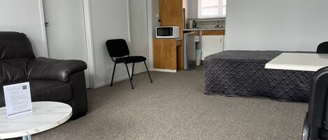 A compact room with a bed, a small table, a chair, a sofa, and a kitchenette in the background.
