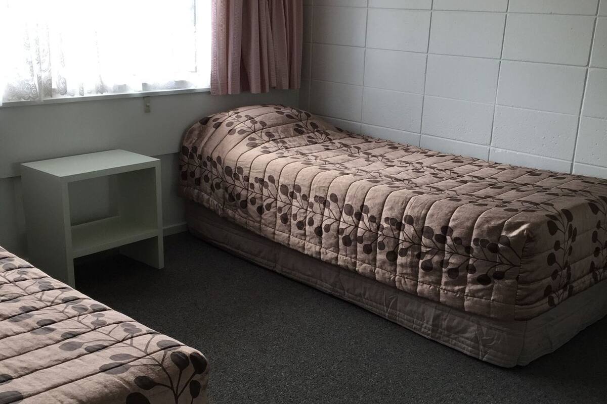 Una cama individual con una colcha de estampado de hojas, una mesita de noche pequeña y blanca, y una ventana con cortinas.
