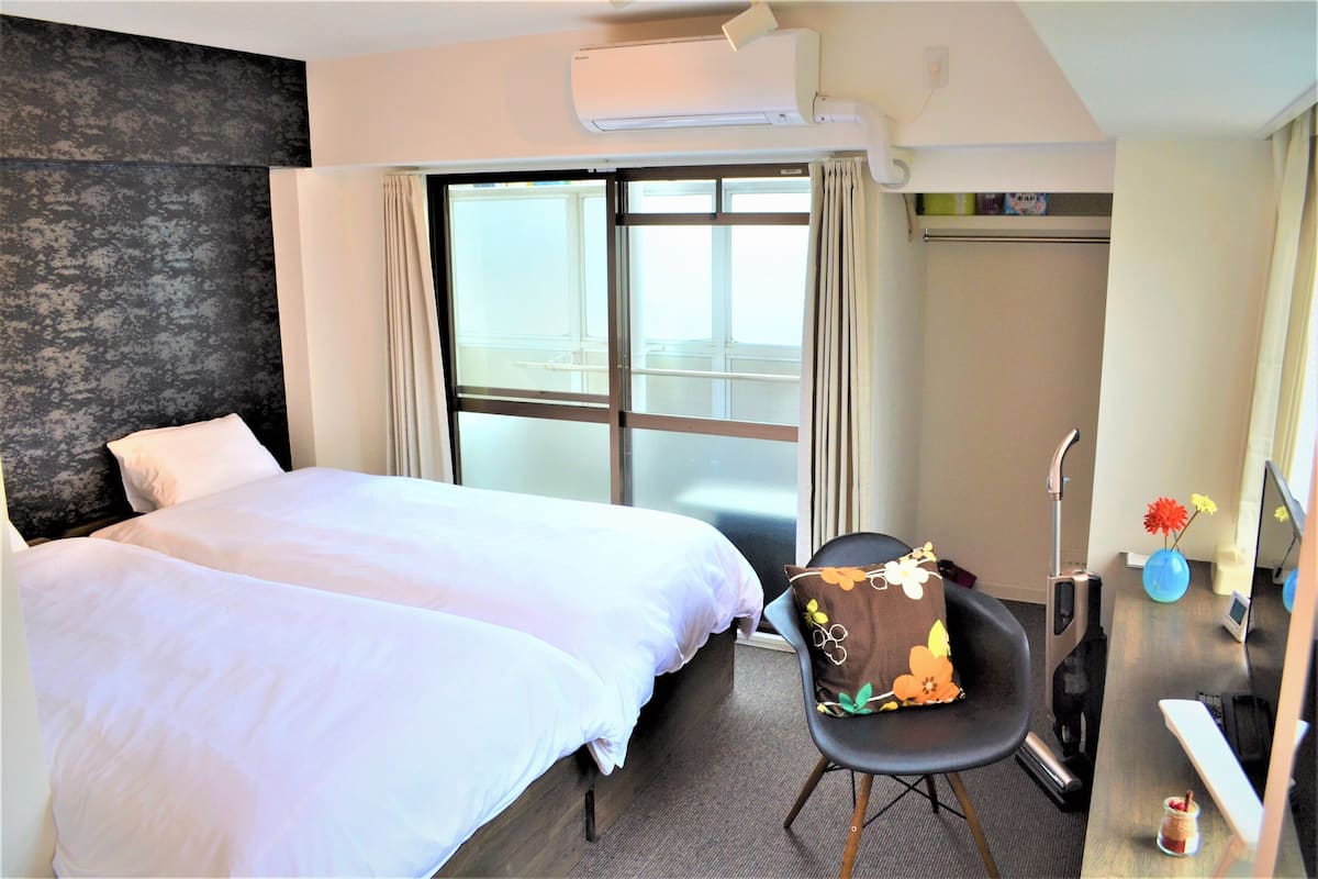 A hotel room with two beds, a desk with a chair, a large window, and an air conditioning unit.