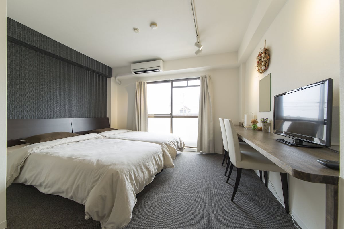 A hotel room with two beds, a desk with a television, and an air conditioning unit.