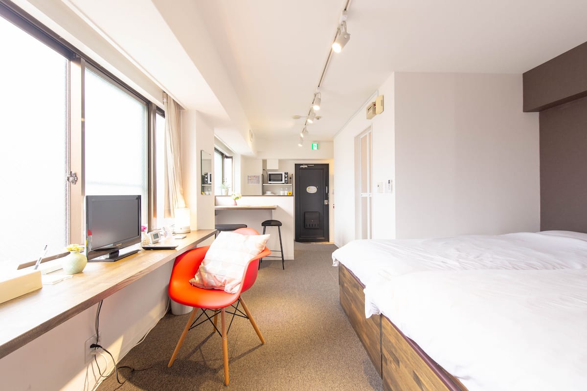 A hotel room with a bed, a desk with a computer, a chair, and a kitchenette in the background.