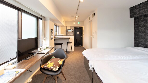 Ett hotellrum med en säng, ett skrivbord, en stol, en TV och ett litet kök.