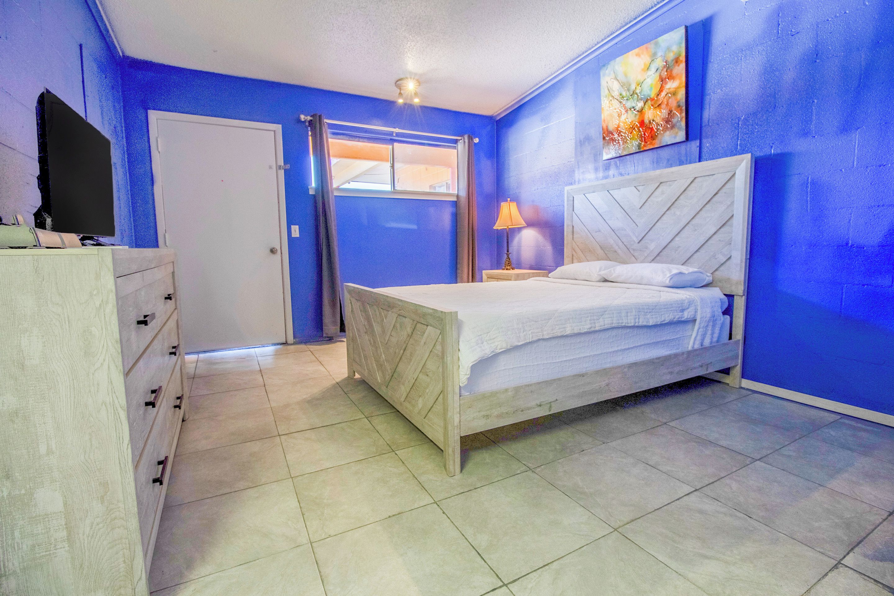 A bedroom with a bed, a dresser, a window with curtains, and a painting on the wall.
