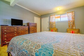 Een slaapkamer met een groot bed, een dressoir met meerdere lades, een televisie erboven en een raam met patroon gordijnen.