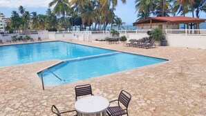 A swimming pool with lounge chairs and a table, surrounded by palm trees and a view of the ocean.